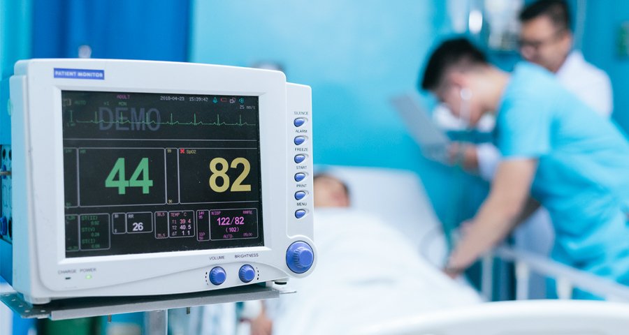 Patient monitor displaying vital signs in an ICU room with healthcare professionals attending a patient in the background.